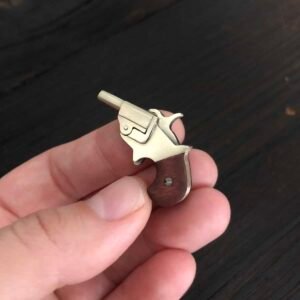 A hand is holding Product Name 1, a miniature brass and wood revolver that resembles a keychain or mini firearm. The background is a dark wooden surface. This tiny piece is intricately detailed, featuring a visible trigger and hammer.